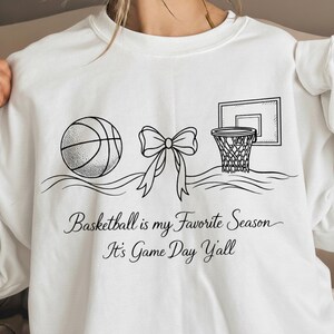 May include: White sweatshirt with a black line drawing of a basketball, a bow, and a basketball hoop. The text "Basketball is my Favorite Season, It's Game Day Y'all" is written in a script font.