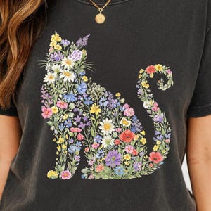 May include: Black t-shirt featuring a floral cat design. The cat silhouette is filled with colorful flowers, including daisies, poppies, and lavender. The shirt has a crew neck and short sleeves, and is made of a soft, dark fabric.