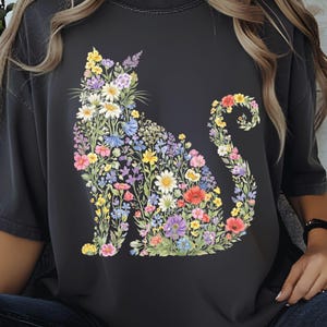 May include: Dark gray t-shirt featuring a floral cat design. The cat silhouette is filled with colorful flowers in shades of yellow, pink, purple, and blue. The shirt has a relaxed fit and short sleeves, perfect for casual wear.