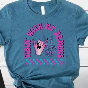May include: A teal t-shirt featuring a graphic design with the text "Vibin' With My Demons". The design includes a hand with black nails and a skeleton hand making a peace sign, with pink and blue accents.