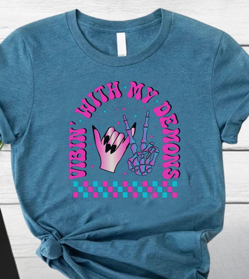 May include: A teal t-shirt featuring a graphic design with the text "Vibin' With My Demons". The design includes a hand with black nails and a skeleton hand making a peace sign, with pink and blue accents.