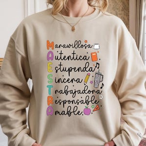 May include: Beige sweatshirt with the word "MAESTRA" in colorful letters, each letter representing a positive adjective. The design includes small illustrations of a book, pencil, and apple. A perfect gift for teachers.