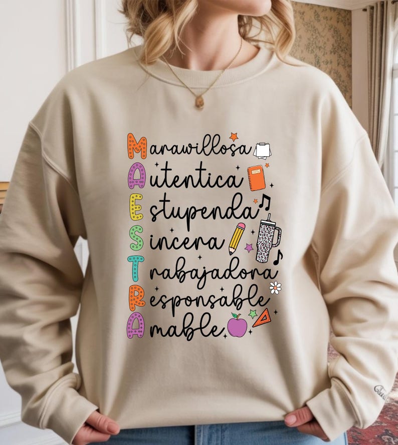 May include: Beige sweatshirt with the word "MAESTRA" in colorful letters, each letter representing a positive adjective. The design includes small illustrations of a book, pencil, and apple. A perfect gift for teachers.