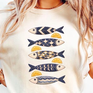 May include: Cream-colored t-shirt featuring five stylized fish designs in navy blue with various patterns. Each fish is paired with a yellow lemon slice. The fish designs include chevron, scale, and wave patterns. A casual, nautical-themed top.