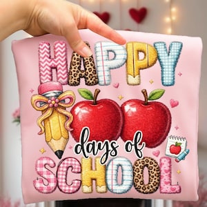 May include: A pink pillow with the words "HAPPY days of SCHOOL" in various patterns and colors. The design includes two red apples, a pencil, and a small notepad with a red apple and pencil illustration. The pillow is held by a hand.