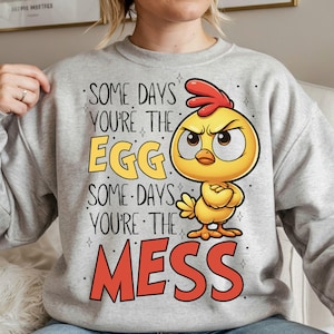 K&ouml;nnte beinhalten: Ein hellgraues Sweatshirt mit dem Text "SOME DAYS YOU'RE THE EGG SOME DAYS YOU'RE THE MESS" und einer Cartoon-K&uuml;ken-Illustration. Das K&uuml;ken ist gelb mit rotem Kamm und hat einen w&uuml;tenden Gesichtsausdruck.