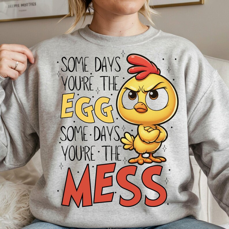 K&ouml;nnte beinhalten: Ein hellgraues Sweatshirt mit dem Text "SOME DAYS YOU'RE THE EGG SOME DAYS YOU'RE THE MESS" und einer Cartoon-K&uuml;ken-Illustration. Das K&uuml;ken ist gelb mit rotem Kamm und hat einen w&uuml;tenden Gesichtsausdruck.