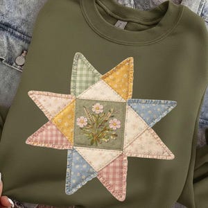 May include: Olive green sweatshirt featuring a star-shaped appliqu&eacute;. The appliqu&eacute; is made of various patterned fabrics, including floral, gingham, and dotted designs, surrounding a central square with embroidered flowers. The sweatshirt has a crew neck and long sleeves.
