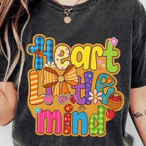 May include: Dark gray t-shirt with a colorful graphic design. The design features the words "Heart Little Mind" in various fonts and colors, along with illustrations of flowers, a heart, a brain, and a basket of apples.