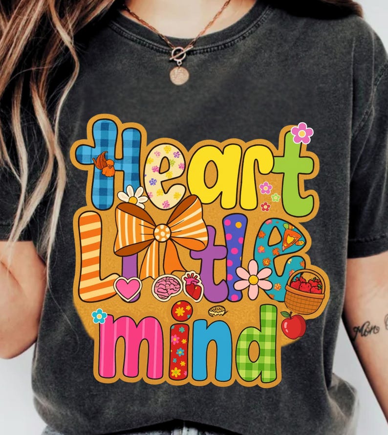 May include: Dark gray t-shirt with a colorful graphic design. The design features the words "Heart Little Mind" in various fonts and colors, along with illustrations of flowers, a heart, a brain, and a basket of apples.