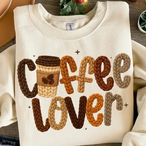May include: Cream-colored sweatshirt with the words "COFFEE LOVER" in a textured, 3D design. The word "COFFEE" features a coffee cup graphic. The letters are in shades of brown, tan, and orange.