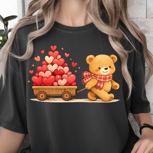May include: A black t-shirt with a cartoon teddy bear pulling a wooden cart filled with heart-shaped balloons. The bear wears a red and white plaid scarf. The cart is filled with red and pink hearts.