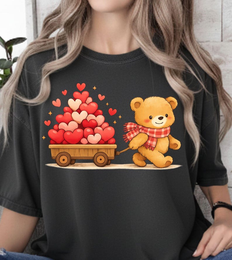 May include: A black t-shirt with a cartoon teddy bear pulling a wooden cart filled with heart-shaped balloons. The bear wears a red and white plaid scarf. The cart is filled with red and pink hearts.