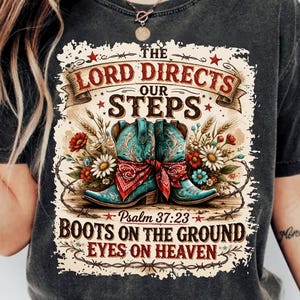 May include: Dark gray t-shirt featuring a graphic with turquoise cowboy boots, a red bandana, and floral accents. The text reads "The Lord Directs Our Steps" and "Boots on the Ground, Eyes on Heaven" with a Bible verse reference.