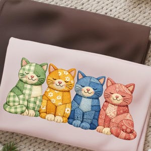 Puede incluir: Una manta rosa claro con cuatro figuras de gato bordadas en verde, amarillo, azul y rojo. Cada gato tiene un diseño de patchwork único y los ojos cerrados, lo que sugiere sueño. La manta está doblada, con tela marrón visible en el fondo.