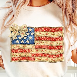 May include: Cream-colored t-shirt featuring a decorative American flag design. The flag is made of fabric with floral and gingham patterns, with a bow at the top. The flag has blue stars and red and white stripes.