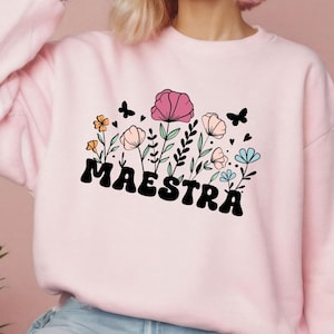 May include: Light pink sweatshirt with the word "MAESTRA" in black, stylized font. The design includes colorful flowers, butterflies, and hearts. The flowers are in shades of pink, orange, and blue.