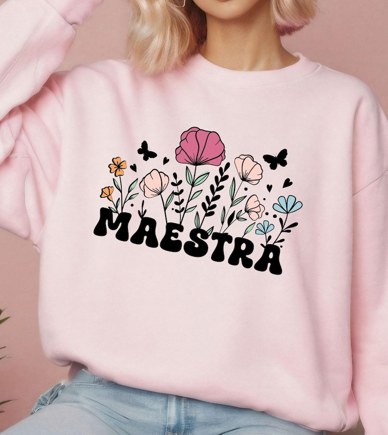 May include: Light pink sweatshirt with the word "MAESTRA" in black, stylized font. The design includes colorful flowers, butterflies, and hearts. The flowers are in shades of pink, orange, and blue.
