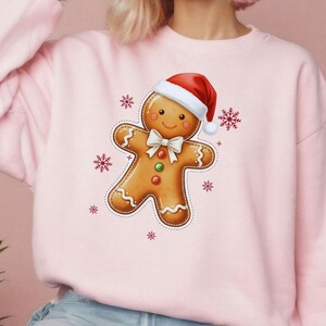 May include: A light pink sweatshirt featuring a gingerbread man graphic wearing a Santa hat and bow tie. The gingerbread man has a smiling face and is surrounded by red snowflakes. A festive holiday-themed sweatshirt.