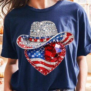 May include: Navy blue t-shirt featuring a patriotic design. The graphic includes a bedazzled cowboy hat with red, white, and blue accents, and a heart-shaped American flag with a large red gem in the center. The shirt has short sleeves.