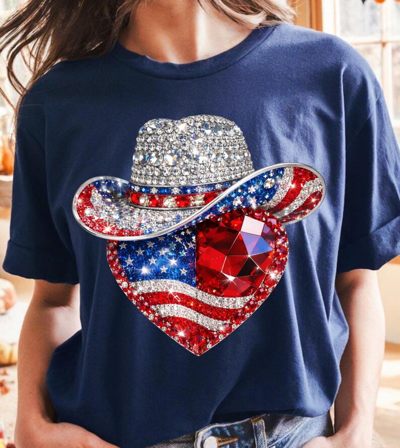 May include: Navy blue t-shirt featuring a patriotic design. The graphic includes a bedazzled cowboy hat with red, white, and blue accents, and a heart-shaped American flag with a large red gem in the center. The shirt has short sleeves.