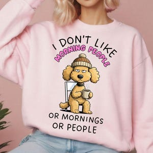 May include: Pale pink sweatshirt with the slogan "I DON'T LIKE MORNING PEOPLE OR MORNINGS OR PEOPLE." The design shows a cartoon dog in a beanie, holding a coffee cup, with a grumpy expression.