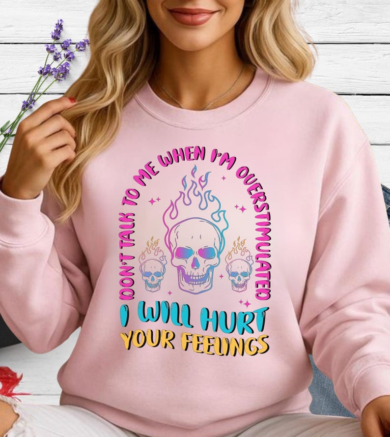 May include: A light pink sweatshirt with a graphic design featuring three skulls and flames. The text reads "DON'T TALK TO ME WHEN I'M OVERSTIMULATED" and "I WILL HURT YOUR FEELINGS" in a colorful, eye-catching font.