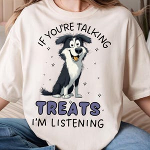May include: A cream-coloured t-shirt with a cartoon dog illustration. The dog has black and white fur and a smiling face. The text reads "IF YOU'RE TALKING TREATS I'M LISTENING" in dark blue, above and below the dog illustration.