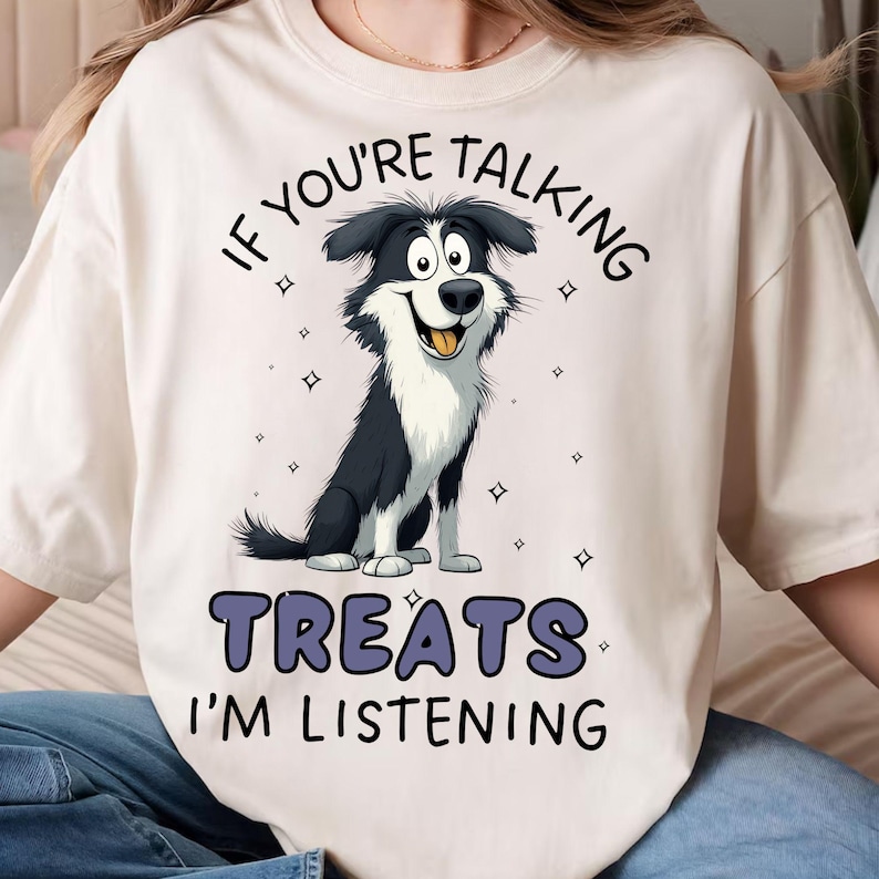 May include: A cream-coloured t-shirt with a cartoon dog illustration. The dog has black and white fur and a smiling face. The text reads "IF YOU'RE TALKING TREATS I'M LISTENING" in dark blue, above and below the dog illustration.