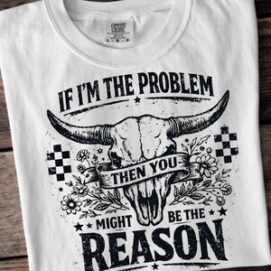 May include: White t-shirt with a graphic design featuring a bull skull, flowers, and the text "IF I'M THE PROBLEM THEN YOU MIGHT BE THE REASON." The shirt has a crew neck and short sleeves. The Comfort Colors tag is visible.
