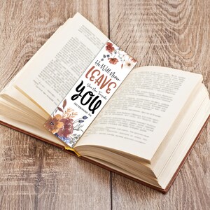 May include: A bookmark with floral accents and the text "He Will Never Leave You" in brown and black. The bookmark is placed in an open book with a wooden background. The text also includes "Deuteronomy 31:6".