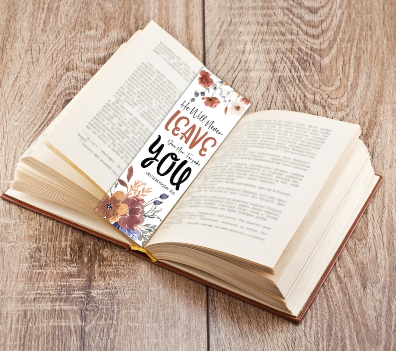 May include: A bookmark with floral accents and the text "He Will Never Leave You" in brown and black. The bookmark is placed in an open book with a wooden background. The text also includes "Deuteronomy 31:6".
