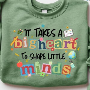 May include: Sage green sweatshirt with the text "IT TAKES A big heart To SHAPE LITTLE minds" in colorful, playful lettering. The design includes school-themed illustrations like a globe, apple, and pencil, perfect for teachers.