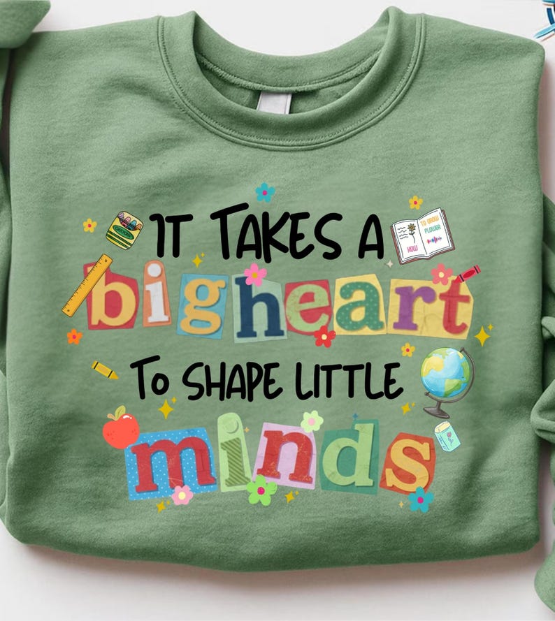 May include: Sage green sweatshirt with the text "IT TAKES A big heart To SHAPE LITTLE minds" in colorful, playful lettering. The design includes school-themed illustrations like a globe, apple, and pencil, perfect for teachers.