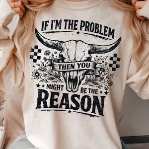 May include: Cream-colored long-sleeve shirt with a graphic of a bull skull, flowers, and the text "IF I'M THE PROBLEM THEN YOU MIGHT BE THE REASON" in black. The design includes checkered patterns.