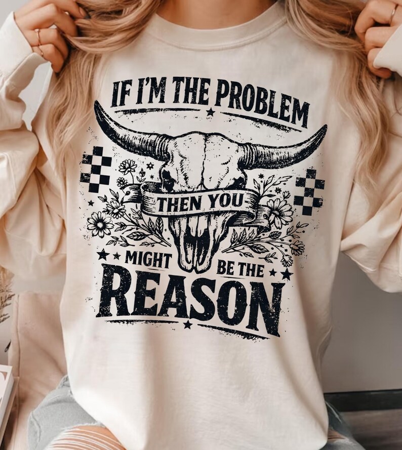 May include: Cream-colored long-sleeve shirt with a graphic of a bull skull, flowers, and the text "IF I'M THE PROBLEM THEN YOU MIGHT BE THE REASON" in black. The design includes checkered patterns.