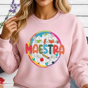 May include: A light pink sweatshirt with a colorful graphic that says "MAESTRA". The graphic is a circle with a rainbow border, featuring stars, flowers, a pencil, an apple, and a book.