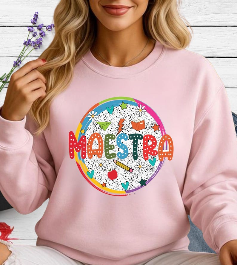 May include: A light pink sweatshirt with a colorful graphic that says "MAESTRA". The graphic is a circle with a rainbow border, featuring stars, flowers, a pencil, an apple, and a book.