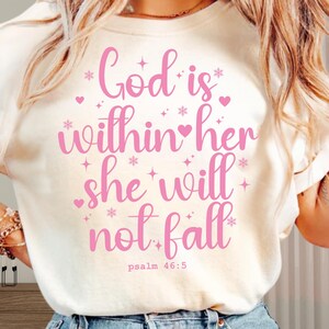 May include: Cream-colored t-shirt with the text "God is within her she will not fall" in pink script. The design includes hearts, stars, and the Bible verse Psalm 46:5.