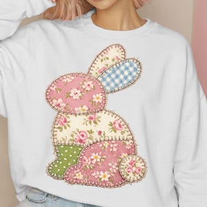 Könnte beinhalten: Weißes Sweatshirt mit einem Patchwork-Hasen-Design. Der Hase besteht aus verschiedenen floralen und gemusterten Stoffen in Rosa, Grün und Blau. Das Design ist mit braunem Faden genäht und wirkt rustikal und handgefertigt.