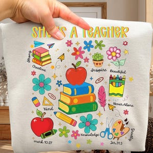 May include: A light beige sweatshirt with the text "She's a Teacher" in colorful letters. The design includes stacks of books, apples, flowers, and pencils. Additional text includes "Strong", "Creative", "Kind", "Inspire", "Beautiful", "Knowledge", "Never Alone", "Greatly", and Bible verses.