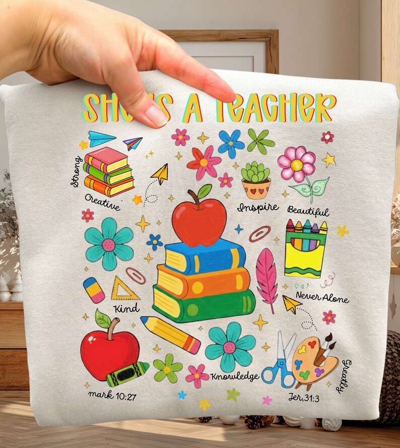 May include: A light beige sweatshirt with the text "She's a Teacher" in colorful letters. The design includes stacks of books, apples, flowers, and pencils. Additional text includes "Strong", "Creative", "Kind", "Inspire", "Beautiful", "Knowledge", "Never Alone", "Greatly", and Bible verses.