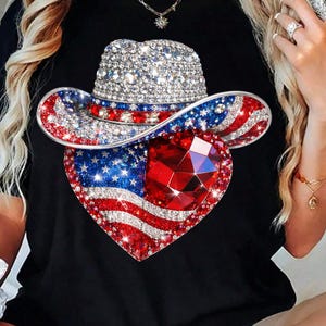 May include: Black t-shirt featuring a patriotic design. The design includes a bedazzled cowboy hat and a heart-shaped emblem with the American flag and a large red gem. The hat and heart are covered in sparkling rhinestones.