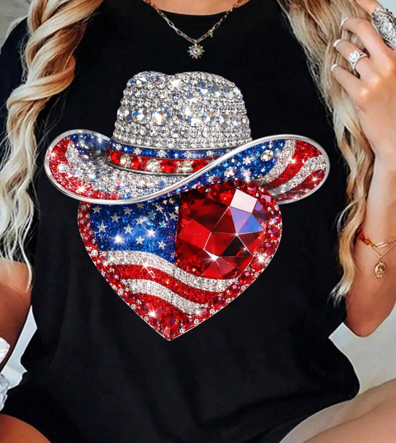 May include: Black t-shirt featuring a patriotic design. The design includes a bedazzled cowboy hat and a heart-shaped emblem with the American flag and a large red gem. The hat and heart are covered in sparkling rhinestones.