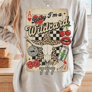 May include: A light gray long-sleeve shirt featuring a graphic with a vintage playing card design. The design includes a bull skull, cherries, dice, and the text "Baby I'm a Wildcard" with the number "777".