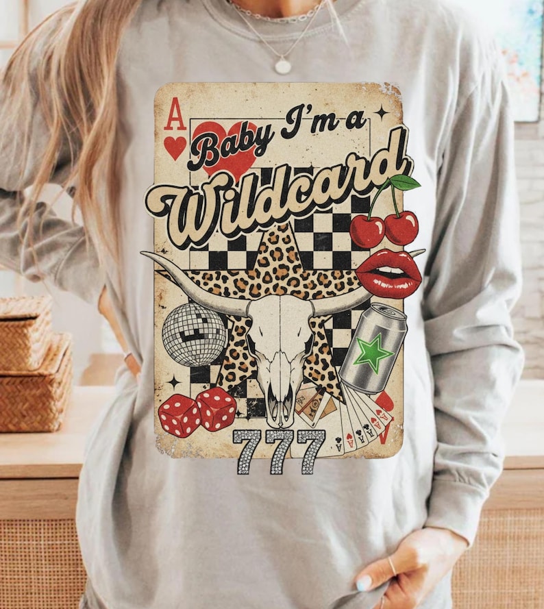 May include: A light gray long-sleeve shirt featuring a graphic with a vintage playing card design. The design includes a bull skull, cherries, dice, and the text "Baby I'm a Wildcard" with the number "777".