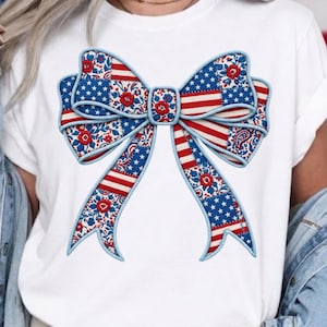 Könnte beinhalten: Weißes T-Shirt mit einem großen Schleifendesign. Die Schleife zeigt patriotische Muster: rot-weiße Streifen, blaue Sternenfelder und florale und Paisley-Akzente. Die Schleife ist mit einer blauen, seilartigen Umrandung versehen.