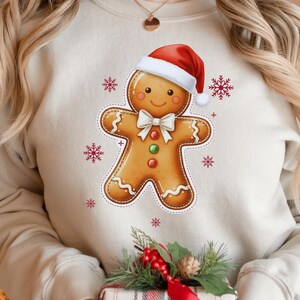 May include: A beige sweatshirt featuring a gingerbread man design wearing a red Santa hat and a white bow tie. The gingerbread man has a smiling face and is decorated with red, green, and white accents. Red snowflakes surround the gingerbread man.