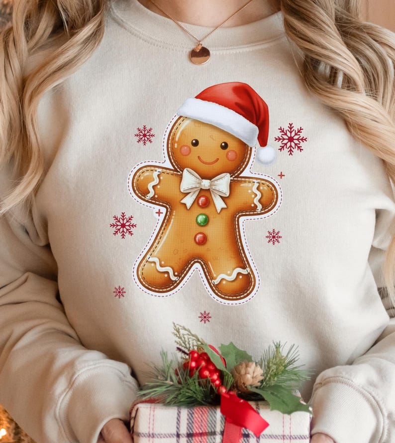 May include: A beige sweatshirt featuring a gingerbread man design wearing a red Santa hat and a white bow tie. The gingerbread man has a smiling face and is decorated with red, green, and white accents. Red snowflakes surround the gingerbread man.