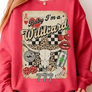 May include: Red sweatshirt with a graphic design featuring a bull skull, cherries, dice, a disco ball, and the text "Baby I'm a Wildcard". The design includes a leopard print star and a can with a green star.